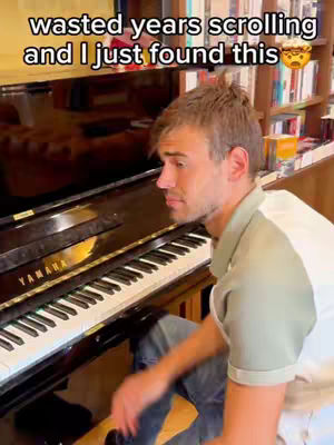 A man sits at a piano, partially facing the camera. He’s wearing a casual green shirt and jeans. The background includes a piano, a wooden shelf with books and objects, and a blurred reflection in the piano's surface.