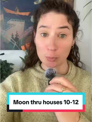 A woman is centered in the frame, looking toward the viewer and holding a microphone. She is wearing a light green sweater and appears to be in her home, with a painting on the wall behind her. A colorful text overlay at the bottom reads, "Moon thru houses 10-12."