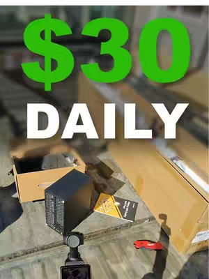 The cover image shows a top-down view of a surface with several objects: a small black computer-like device, a folded yellow paper publication, an opened cardboard box, a red toy car, and a video camera attached to a gimbal. Large green text overlays the top of the image, the words '$30 DAILY' in a bold, white font below it. The background has unclear details, and the objects are scattered, suggesting a workspace or setup for the video.