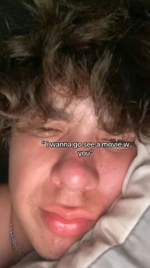 A close-up image of a young man's face. His right eye is slightly visible, and he appears to be lying down, possibly in bed, with a pillow behind him. His hair is dark and curly. The man's lips are prominent, and a silver chain is visible around his neck. Text overlayed on the image reads, "i wanna go see a movie w you".