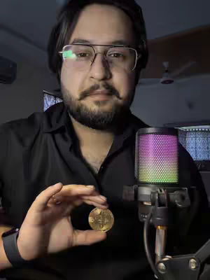The cover image shows a man in a black shirt, holding a Bitcoin coin in his right hand. He is wearing glasses and has a beard and mustache. A microphone on a stand is to his right, with a colorful light display. The background is a room with a ceiling fan.