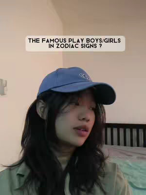 The image shows a young woman with long black hair and a blue baseball cap, facing slightly to her right. Above her head, a light-colored rectangle contains the text: 'THE FAMOUS PLAY BOYS/GIRLS IN ZODIAC SIGNS ?' The background is simple, with cream-colored walls.