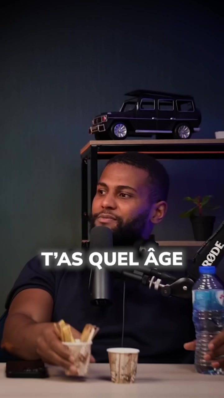 A man is seated in a studio-like setting, mid-chest up, with a microphone in front of him. Text overlayed on the image asks "T'AS QUEL ÂGE". Beside him, a bottle of water and containers with snacks sit on a table. Behind him is a black car model on a shelf.