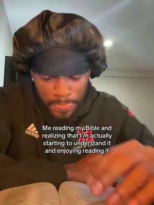 A man wearing a black durag and a dark green hoodie is seated, reading a book that is open in his hands. The text overlay reads, 'Me reading my Bible and realizing that I'm actually starting to understand it and enjoying reading it.' The background includes a hint of a light source and the upper corner of a wall.