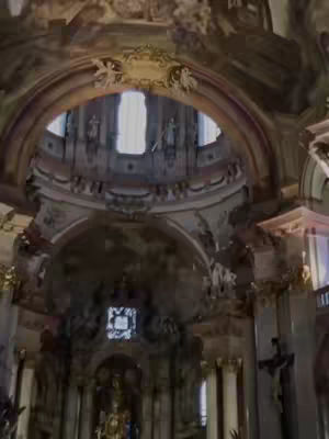 The image captures the interior of a grand church. It features a high, arched ceiling with intricate architectural details and ornate decorations. The space is well-lit, with light streaming in through a central opening, illuminating the details of the arches and the decorative elements. The composition emphasizes symmetry and the impressive scale of the building.