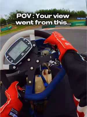 The cover image is a POV shot from inside a go-kart. The foreground shows the steering wheel with a digital display, the driver's red-gloved hands, and splashes of blue. The background features a race track with green barriers and some trees under an overcast sky.