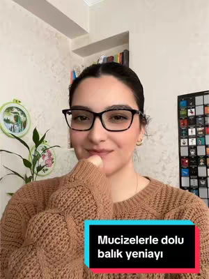A woman with dark hair and glasses is smiling, looking directly at the camera. She is wearing a tan sweater, and a text overlay in a dark-blue rectangle. The background includes a wall with a floral pattern and a side table with a plant and decorative items.