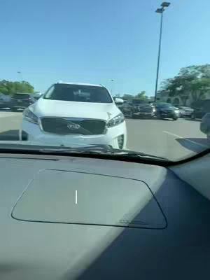 The image offers an eye-level point-of-view shot from inside a car. The camera is pointed towards the front, with the dashboard and windshield visible. Outside, a sunlit parking lot is partially blocked by other vehicles and trees. Dominant colors include the car's interior tones and the blue sky, creating a simple, everyday scene.