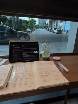 A well-lit, indoor shot of a study desk against a large window. A laptop displaying a black and white program, an open notebook with writing, a drink in a plastic cup, and a pink item are on the desk. Outside, cars are parked on a tree-lined street. The scene presents a study setup with a view.