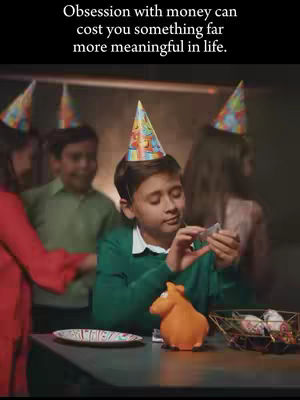 The image shows a group of children at a party. One boy is the focus, holding money while wearing a party hat. Other children are in the background, also wearing party hats. There's a table with a hippo figurine, a plate, and a basket with food. The background appears dark.
