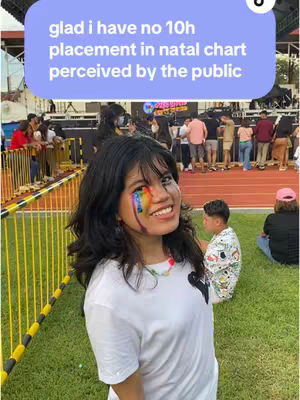A woman with dark hair and a rainbow eye design smiles in front of a grassy area crowded with people in an outdoor setting. A text overlay in a light blue bubble says "glad i have no 10h placement in natal chart perceived by the public",