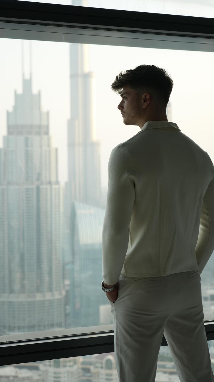 A man is viewed from the back and slightly to the side, gazing out a large window at a cityscape. The man is wearing a light-colored sweater and pants, and his watch is visible on his wrist. The cityscape in the background includes several tall buildings, suggesting a modern urban environment. The overall composition is clean and the lighting is soft.