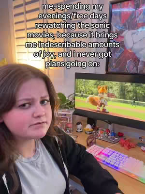 A young woman with brown hair is seated, looking at the camera and appearing slightly exasperated. Behind her, a computer monitor displays a scene from an animated movie, featuring a cartoon fox character. Additional elements include a keyboard, desk ornaments, and window blinds, suggesting a home office or entertainment setup.