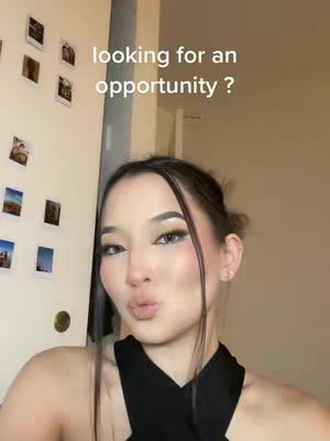 A selfie-style image with a young woman in the foreground. The woman has long dark hair with a middle part, elegantly styled and pulled back. She wears makeup that accentuates her eyes and lips, creating a refined look. Her expression is subtly alluring, lips slightly pursed as if she is about to kiss the camera. Her top is black, contrasting with her light skin tone. Behind her, on a white door, are several framed photos. The background wall is light brown, and some light spills in naturally, forming a soft visual balance.