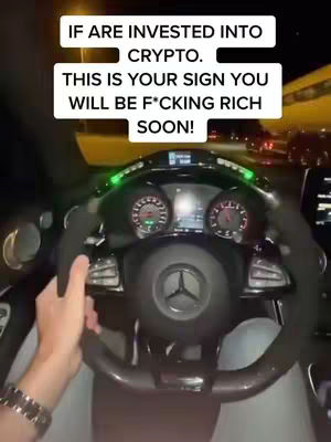 A first-person perspective from the driver's seat of a car at night, with the steering wheel dominating the frame and overlaid text in a rectangle shape, while a person's hand is visible on the steering wheel. Streetlights and other vehicles are visible in the background