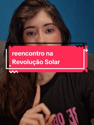 A woman with long brown hair is centered in a selfie-style shot. She is directly facing the camera, with the top of the image cropped at her hairline. She is wearing a black shirt, and her hand is up with her index finger pointing upwards. A rectangular overlay in bright red, white, and black spans across her face, with text that reads "reencontro na Revolução Solar". The background is a blurred blue.