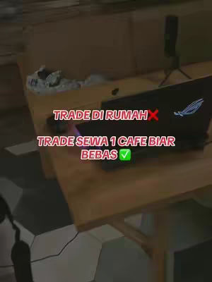 The image shows a laptop open on a wooden table in what appears to be a cafe or co-working space. Text overlays are present, contrasting trading from home with renting a cafe for more freedom and a relaxed trading environment. The overall composition is a bit dark, but the focus seems to be on the laptop and the message.