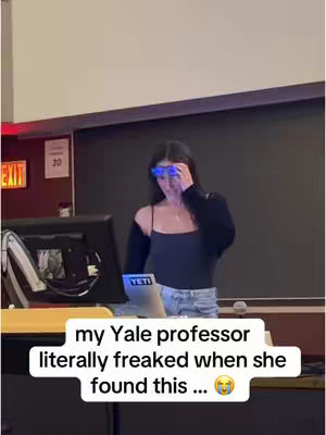 A woman in a black top and jeans is standing in what seems to be a lecture hall, with a laptop on a desk in front of her. She is wearing blue-tinted glasses and is holding her hand up to her face in surprise. The lighting is bright and the overall composition is straightforward, focused on the woman as the central figure.