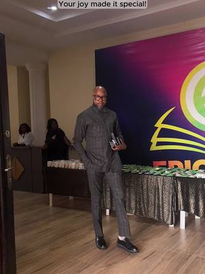 The image captures a medium shot of a well-dressed man standing indoors. He is wearing a dark gray, checkered suit and black shoes, holding an item in his hands. He has a bald head and glasses. Behind him, a large colorful banner, with what appears to be part of a logo on the right.