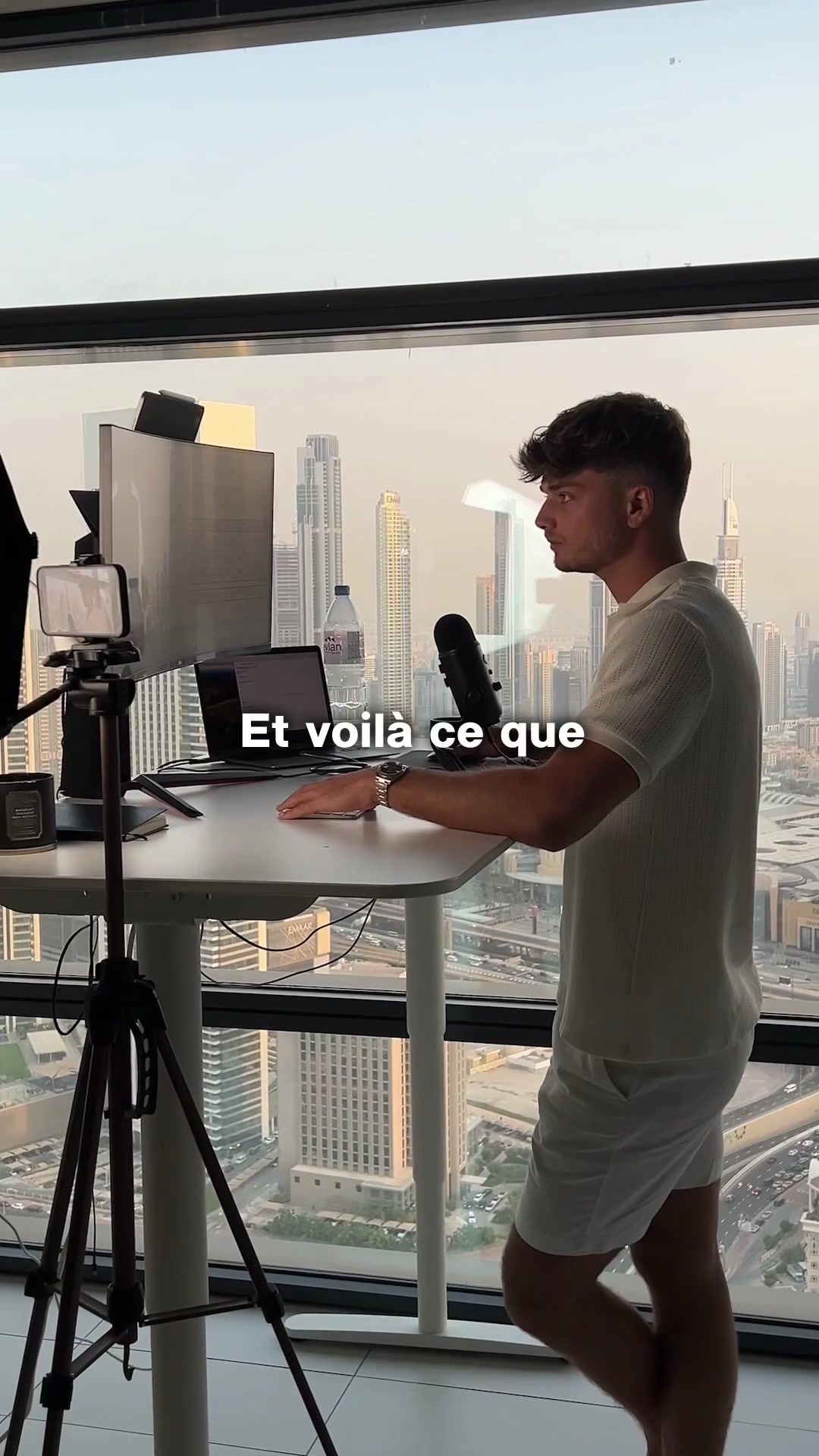 The cover image displays a man standing at a white desk with a cityscape visible through a large window behind him. The scene is brightly lit, presumably by natural light. On the desk are a laptop, microphone, phone, and other recording equipment. The man is wearing a light-colored outfit and looking to the side.
