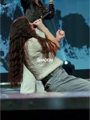A woman with long, wavy brown hair is singing into a microphone. She is wearing a white sweater and blue jeans. The background is a blurry blue and white pattern. The image has a dynamic feel.