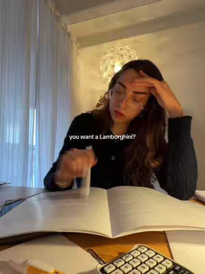 A woman with long brown hair wearing glasses is seated at a table, resting her head on her left hand. She is looking at a notebook open in front of her, with a pen in her right hand and another in her mouth. A calculator is visible, along with a lampshade. The color palette includes neutral tones with some yellow from the lights, along with the dark color of her sweater.