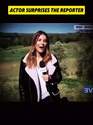 A female reporter stands outside, holding a microphone. The background shows a green field and some trees. The weather looks sunny and bright.