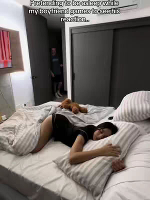 A bedroom scene with a woman lying on a bed, seemingly asleep, with a man visible in the background. The woman is on her side, covered with a blanket, with a pillow under her head. The bed is covered with white sheets and pillows. A brown teddy bear is on the bed. The man is partially visible in the doorway.