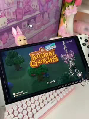 The cover image shows a Nintendo Switch screen displaying the 'Animal Crossing: New Horizons' game, placed on a table in front of a keyboard, controllers, and other pink decor accessories. Text on the screen reads 'Animal Crossing' with the 'New Horizons' subtext, and a button prompt to 'Press +'. The overall aesthetic is bright, cute, and has a cozy atmosphere.