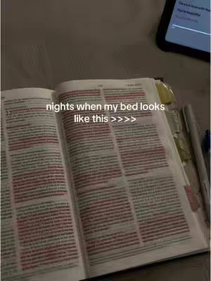 An open Bible lies on a bed or similar surface, the pages spread wide to reveal text. The image is taken from a slightly overhead angle. The lighting is warm-toned. There is a phone in the background that is showing a screen.