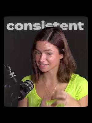 A woman with light skin and brown hair appears on a black background. The image is framed in a portrait style, and the woman is positioned in the center, slightly angled towards the right. She is holding a microphone. Above her head, in large, bold, white letters, the word "consistent" is displayed. The woman is wearing a bright, neon yellow shirt and a delicate gold chain. The overall composition is simple, with the dark background contrasting the bright clothing and text.