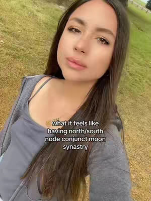 A selfie-style shot featuring a woman looking directly at the camera. She has long, dark hair and is wearing a gray top and jacket. The background is a blurred view of a grassy area with some trees, suggesting an outdoor setting. Text overlayed on the image provides additional context.