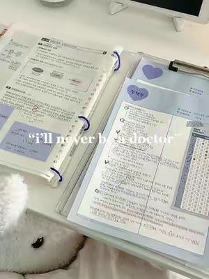 A top-down shot of a desk covered with study materials. There are open notebooks with handwritten notes, a clipboard with papers and a heart-shaped sticker, and a few pens scattered about. The colors are light and airy, with a focus on white, blue, and pale pink.