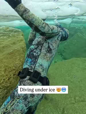 A person in a diving suit is seen submerged in cold water, with a snowy or icy surface above. The person's arm is extended toward the surface. A text overlay says "Diving under ice" with emoticons.