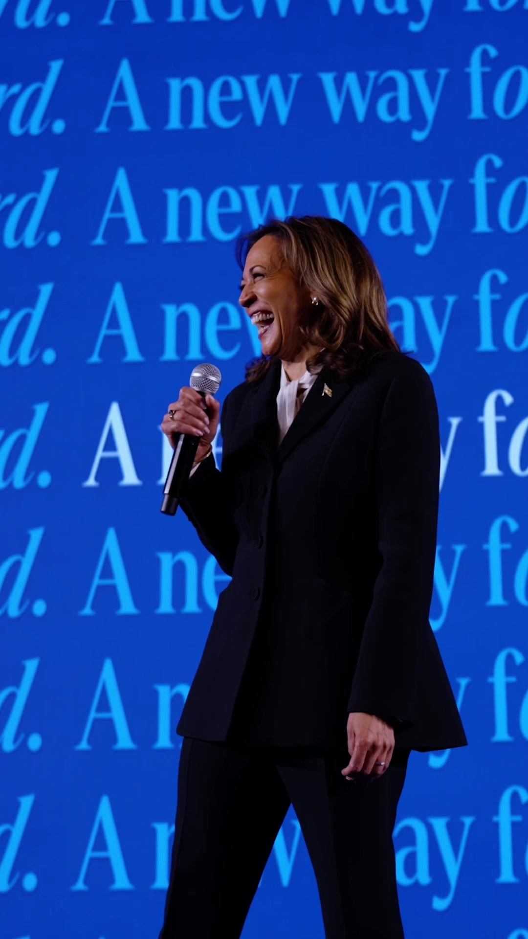 An image of a woman, likely in a professional setting, is captured from the waist up, smiling and holding a microphone. She's wearing a dark suit with a white collared shirt, a small pin and earrings are visible. Behind her is a blue backdrop with white text repeating "rd. A new way fo". The lighting appears to be consistent across the image.