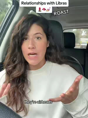 A woman is filming herself inside a car, looking at the camera and speaking. Above her head is text that says "Relationships with Libras ROAST". In the bottom of the image is text that says "they're airheads".