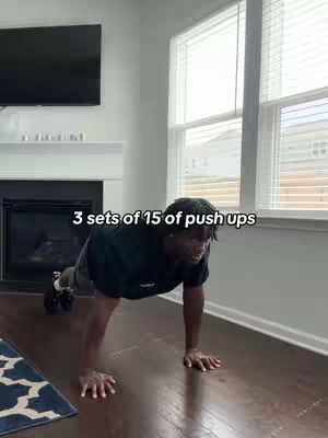 A man is shown in a home interior performing push-ups. The setting includes hardwood floors, and a window allowing natural light to enter. The man is framed centrally while performing the exercise. The text overlay informs the viewer about the sets and reps of push-ups.