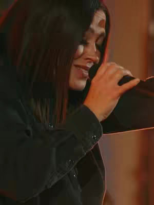 A woman with dark hair, wearing a dark shirt, is holding a microphone and singing. The background is out of focus with warm lighting.