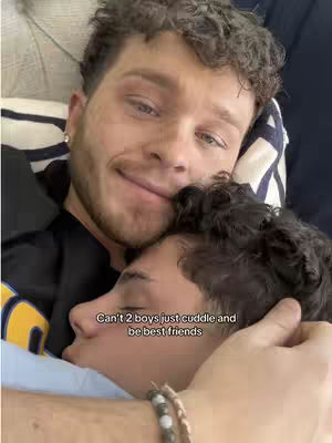 The image shows two men lying in bed, embracing each other. One man has his head resting on the other's chest, who is looking at the camera and smiling. The lighting is soft, enhancing the intimate feel of the scene. The background is slightly blurred, but indicates a bed setting. A text overlay is visible at the bottom: 'Can't 2 boys just cuddle and be best friends'.