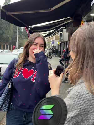 Two women are in an outdoor setting, with one holding a microphone and the other covering her mouth while laughing. The background includes a street with buildings, parked vehicles, and passersby. A coin featuring a Solana logo is in the foreground.