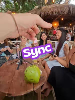 A hand points towards a group of people sitting around a table. An overlay features the word "Sync". A coconut rests on the table.