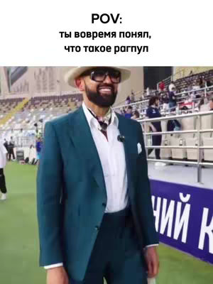 A man in a teal suit and sunglasses stands in a stadium, smiling. The background shows a blurred view of the stadium and people in the stands.