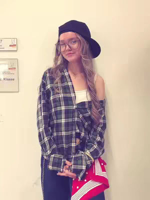 A young woman stands in front of a uniform cream-colored wall, dressed in a layered outfit with a 2000s hip-hop style. She wears a dark baseball cap, large-framed glasses, and a partially unbuttoned flannel shirt over a white top. A red and white patterned bandana is tucked into her dark blue pants pocket.