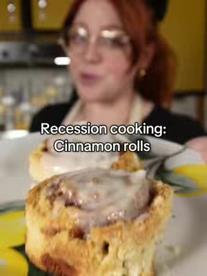 A woman with red hair is holding a plate with cinnamon rolls. The rolls are golden-brown with white icing. The background is blurred, showing a kitchen environment with warm lighting.
