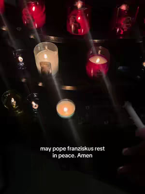 A low-angle shot inside a dimly lit space, likely a church or similar setting, showcasing multiple lit candles. Various sizes of candles in mostly red and white glass holders are arranged on a dark surface, casting warm, flickering light. Beams of light subtly cut through the scene, adding depth and a sense of solemnity. At the bottom, a brief text overlay reads, 'may pope franziskus rest in peace. Amen'.
