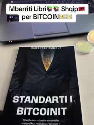 The cover image shows a book titled 'STANDARTI I BITCOINIT' (Bitcoin Standards) placed on a surface with a laptop in the background. The book has a dark cover featuring a person in a suit. Above the book, text overlays state 'Mberriti Libri Shqip per BITCOIN'. The overall composition suggests a theme related to Bitcoin and potentially Albanian language.