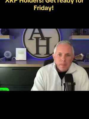A man in a white sweater is seated in front of a desk. Behind him is a lit, circular logo. The man is positioned in the center with a microphone in front of him.