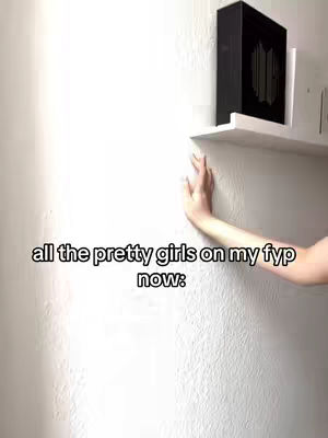 The image shows a white wall with a hand touching it near the bottom right corner. On the upper right, there's a white shelf with a black box on it, likely holding merchandise. Text overlayed in the bottom states 'all the pretty girls on my fyp now:'.