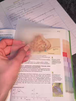A hand holding a clear sheet over an open textbook. The textbook is open to a page with a diagram and text in German. The clear sheet covers part of the diagram.