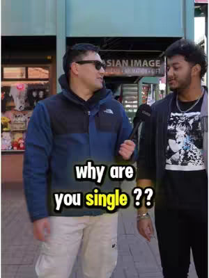 Two men are standing on a city street. The man on the left is wearing a blue jacket, white pants, and sunglasses, and holding a microphone. The man on the right is wearing a black jacket. Text overlay says, "why are you single ??".